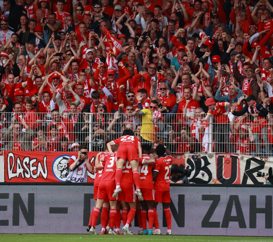 Jogadores do Union Berlin se abraçam, lado a lado, na comemoração de um gol, com torcedores nas arquibancadas ao fundo